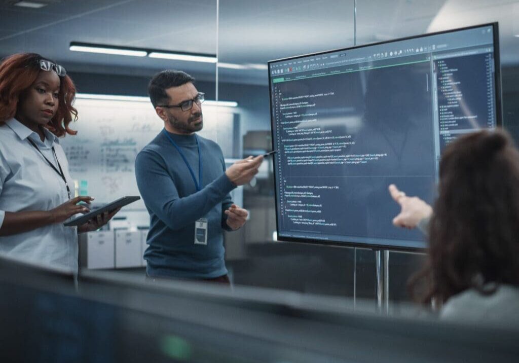 A group of people standing around in front of a monitor.